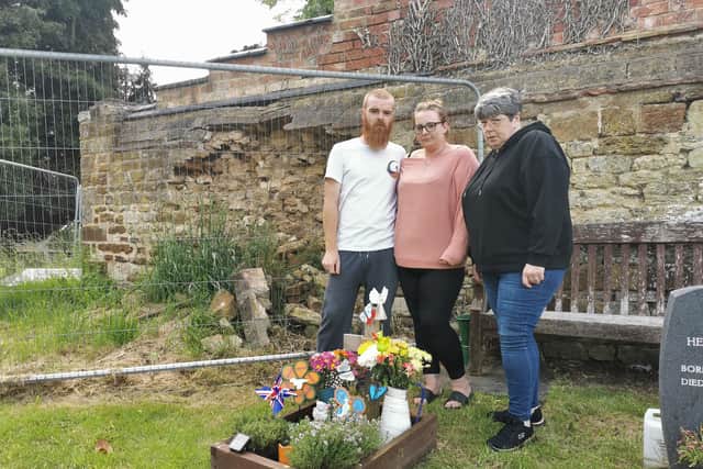 Sharni, her partner and her mother-in-law next to Eddie's grave and the broken wall in the background