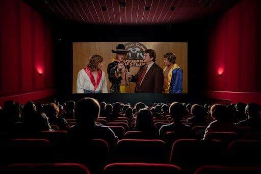 A darkened cinema. A busy audience sits on red velvet seats looking up at the screen. On-screen is a